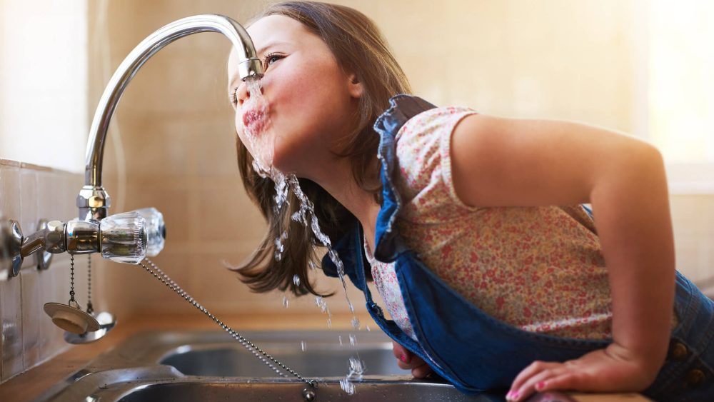 A girl drinking water from a kitchen sink faucet