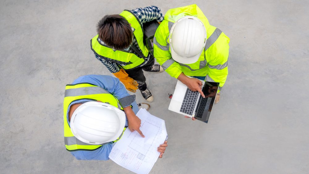 Three workers inspect a blueprint and laptop